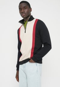 Quarter-zip trui in zwart, crème en rood. Gebreid materiaal met een gestructureerd crème paneel en rode strepen. Voorzien van geribbelde manchetten en zoom.