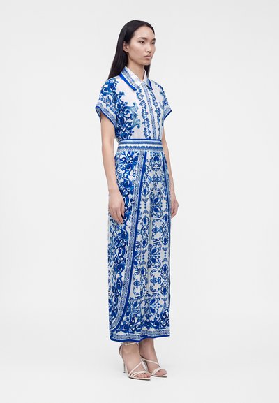 Femme debout portant une robe longue à manches courtes avec col, à motif bleu et blanc, et des sandales blanches à talons hauts sur un fond uni.
