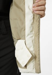 Beige pufferjack interior met een lichtcrème voering, voorzien van een driehoekige zak en vier ronde drukknoopjes. Glad textuur.