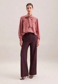 Rosa Satinbluse mit Schleife am Hals, durchknöpfbarem Vorderteil, langen Ärmeln, kombiniert mit tiefburgunderroten, weit geschnittenen Hosen und schwarzen Sandalen.