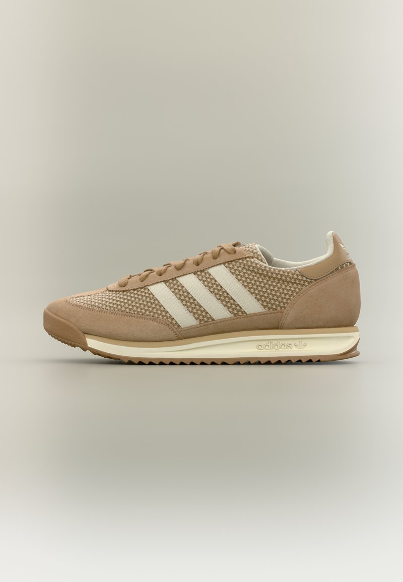 Μπεζ αθλητικό παπούτσι Adidas με λευκές ρίγες, επάνω μέρος από σουέτ και δίχτυ, αναπαυτική σόλα και διακριτικές λεπτομέρειες με το λογότυπο στη φτέρνα και στη μέση της σόλας.