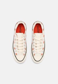 Paire de baskets Converse blanches avec des motifs floraux rouges, des lacets blancs et des semelles intérieures rouges, vues de dessus sur un fond uni.