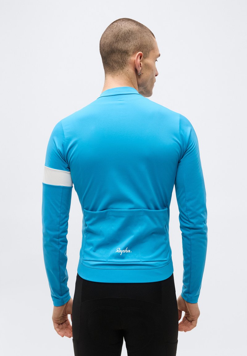 Man facing away wearing bright blue long-sleeve cycling jersey with white armband and "Rapha" logo above rear pockets.