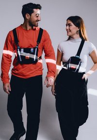 Två individer bär svarta selen-stil väskor med justerbara remmar, som har en blandning av färger och reflexdetaljer över avslappnade kläder.