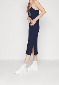 Žena v námořnickém modrém midi šatech s bílými ramínky a bočním rozпарším, doplněných o bílé ponožky a masivní bílé tenisky.