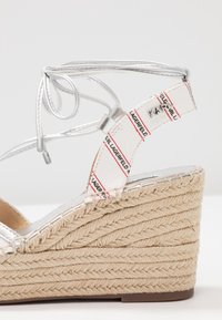 Srebrne sandały z plecionym klinem z juty, z teksturowanym paskiem z wzorem logo i krzyżującym się wiązaniem w okolicy kostki.