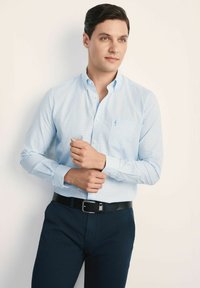 Jasnoniebieska koszula w kratę z guzikiem i logo na kieszeni, zestawiona z ciemnoniebieskimi spodniami i czarnym paskiem. Gładka, bawełniana struktura.