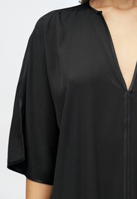 Crna haljina s dubokim V-izrezom, polu-rukavima i glatkim, strukturiranim materijalom. Ima minimalistički dizajn s suptilnim detaljima šivanja.
