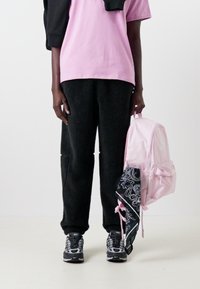 Svarta fleecebyxor, rosa oversized t-shirt, svarta sneakers och en ljusrosa ryggsäck med en mönstrad scarf fäst på ena sidan.