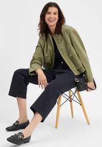 Olijfgroene jas met een bruine kraag, zwarte broek en zwarte loafers. Kleine handtas met gouden accenten, zittend op een houten stoel.