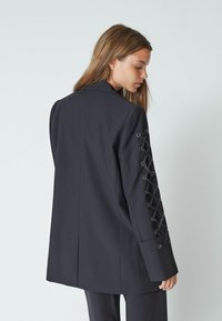 Črni blazer z oblikovanim krojem in vezanjem na levem rokavu s črnim strojčkom. Gladko blago, srednje dolžine.