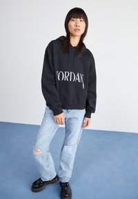 Sudadera negra con el logotipo "JORDAN", corte holgado, capucha con cordón, combinada con pantalones vaqueros desgastados de color azul claro y zapatillas de plataforma negras.