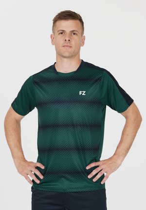 Mies seisoo käsivarret lanteilla, pukeutuneena tummanvihreään ja mustaan kuvioituun urheilut-paitaan, jossa on "FZ"-logo rinnassa.