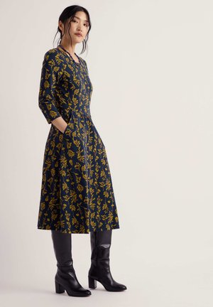 Woman wearing a navy blue knee-length dress with yellow floral pattern and black heeled knee-high boots, standing with hands in pockets.