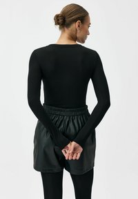 Czarny dopasowany top z długim rękawem, zestawiony z czarnymi skórzanymi szortami z wysokim stanem. Modelka z dłońmi złożonymi za plecami, podkreślono gładkie faktury.