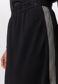 Zwarte rok met een gladde textuur en een elastische tailleband. Voorzien van een zijstreep met dunne, afwisselende zwarte en witte strepen.