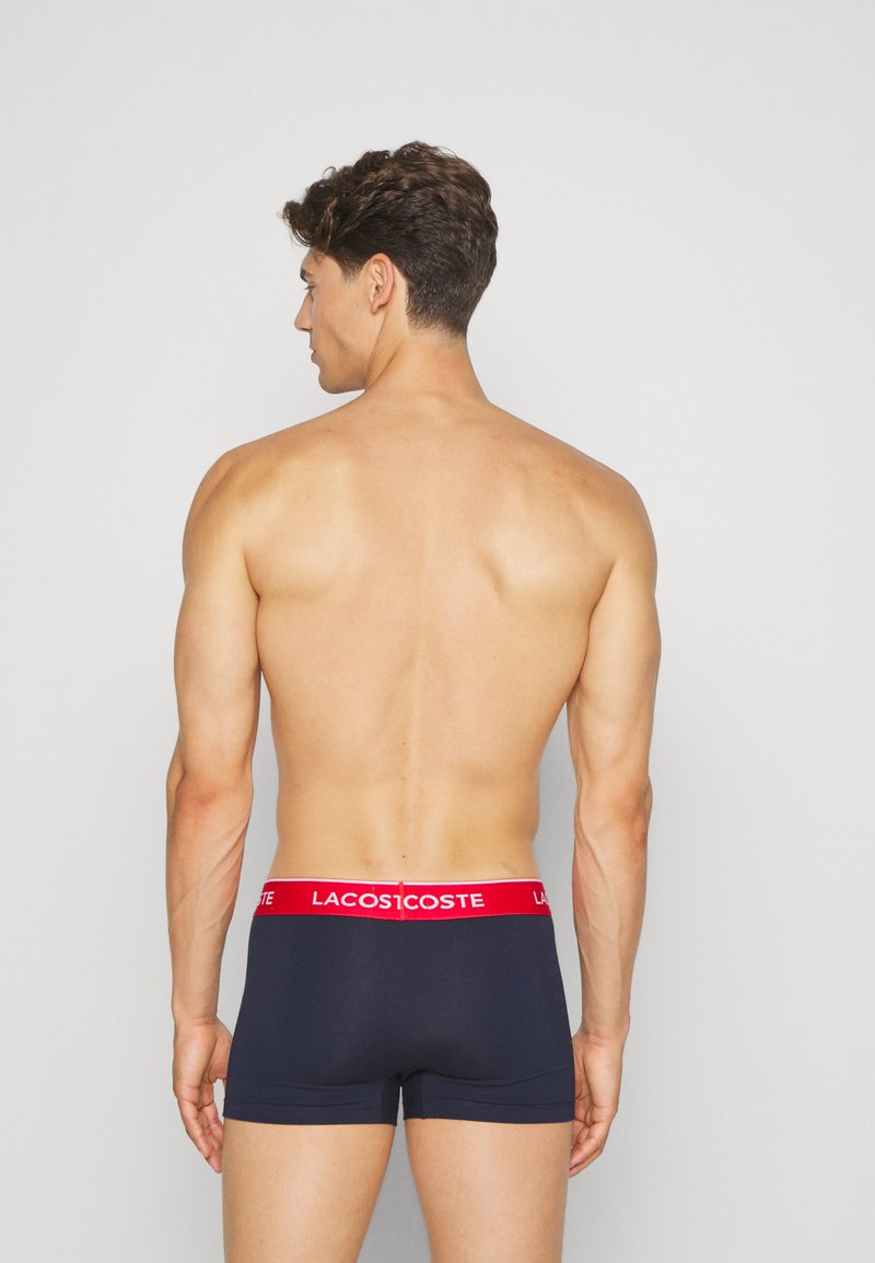 Boxer-briefs marine avec une ceinture rouge arborant le logo Lacoste, présentant une texture lisse et une coupe ajustée. Détails de coutures visibles sur les bords.