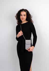 Borsa a tracolla grigia in materiale morbido e texturizzato, con chiusura a patta. Indossata sopra un vestito nero a righe. Cinghia regolabile, design slim.