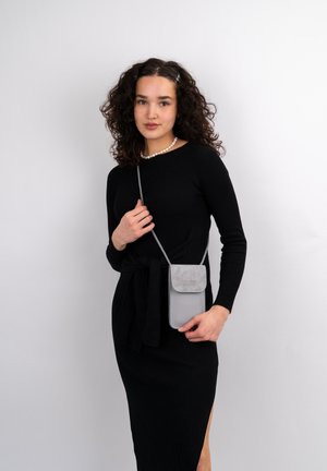Graue Umhängetasche aus strukturiertem, weichem Material mit Klappenverschluss. Über einem schwarzen, gerippten Kleid getragen. Verstellbarer Riemen, schlankes Design.