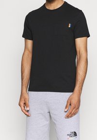 Black cotton t-shirt with a round neck and short sleeves. Features a small multicolored logo patch on the left chest pocket.