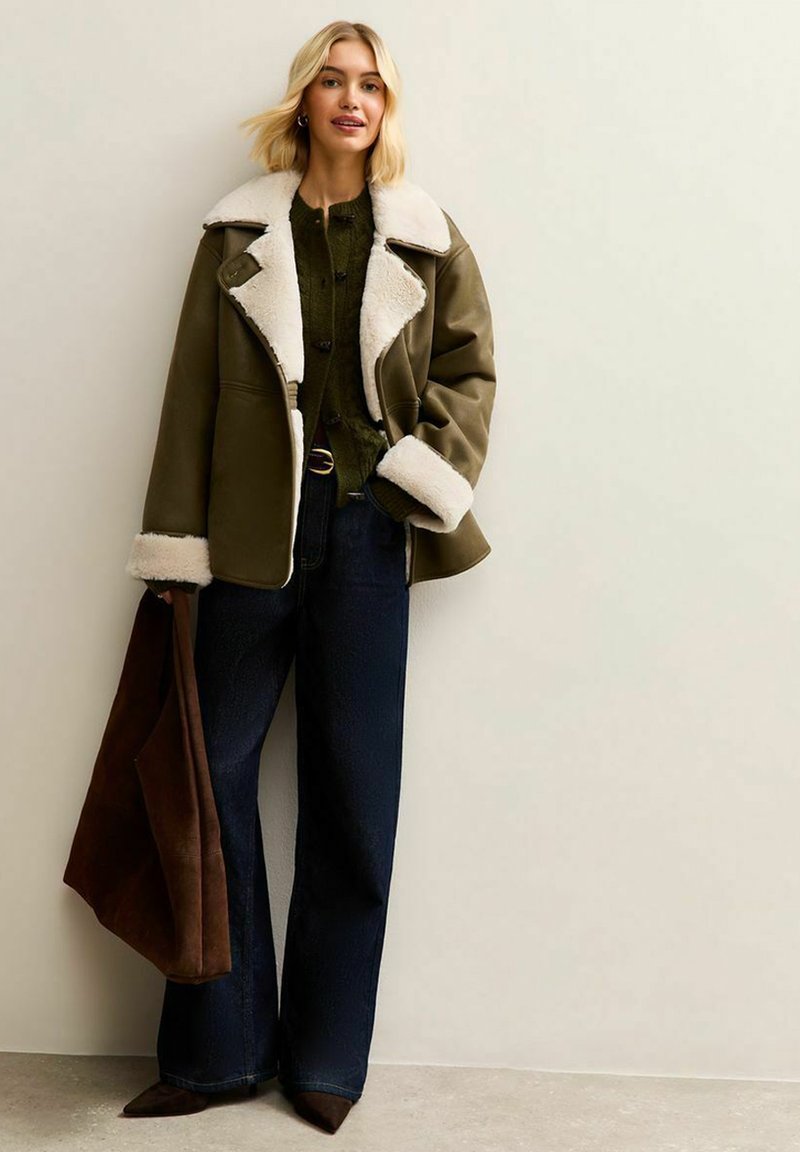 Chaqueta de piel de oveja de color verde oliva forrada de piel de oveja con un cuello amplio, jeans acampanados de color azul oscuro y un bolso de ante marrón. Materiales destacados: cuero, forro de lana.