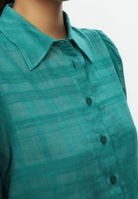 Primer plano de una persona que lleva una camisa de botones verde azulado con cuello puntiagudo y sutiles rayas horizontales texturizadas.