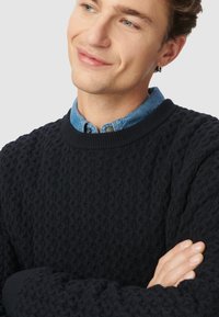 Granatowy sweter wełniany z teksturowanym wzorem, okrągłym dekoltem i ściągaczami. Noszony na jasnej, dżinsowej koszuli, ukazujący stylizację z warstwami.