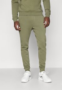 Oliven grønne joggebukser med tapered fit, som har en glatt tekstur, elastisk livlinje og sidelommer. Kombinert med hvite joggesko.