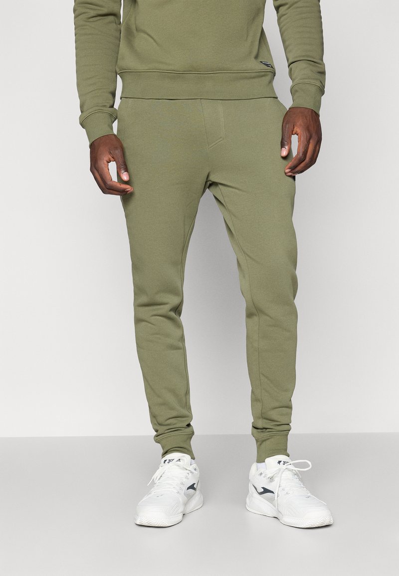 Oliven grønne joggebukser med tapered fit, som har en glatt tekstur, elastisk livlinje og sidelommer. Kombinert med hvite joggesko.