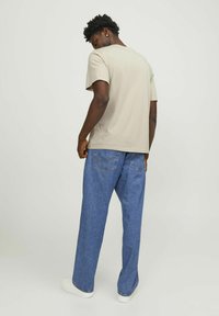 Beige bomull t-skjorte med en avslappet passform, parret med blå denimjeans med baklommer. Modellen står med en lett vridning, iført hvite joggesko.