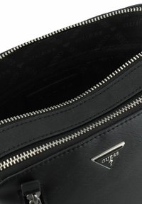 Bolso de cuero negro texturizado con cremallera plateada y logotipo. Interior forrado con un patrón de "GUESS" repetido. Diseño funcional y elegante.