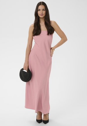 Femme aux longs cheveux bruns portant une robe longue rose sans manches, tenant un sac à main noir tressé et portant des talons noirs ainsi qu'un bracelet en or.