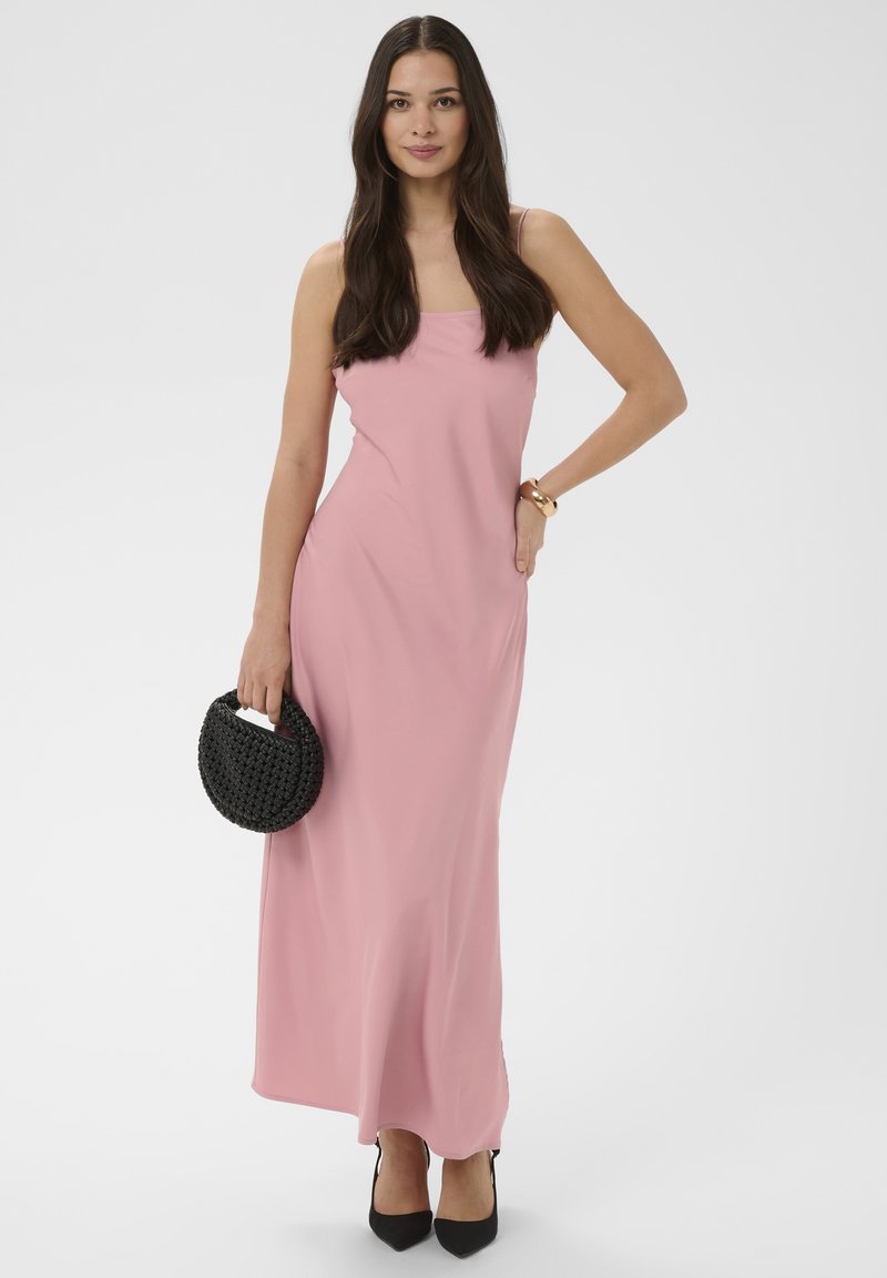 Femme aux longs cheveux bruns portant une robe longue rose sans manches, tenant un sac à main noir tressé et portant des talons noirs ainsi qu'un bracelet en or.