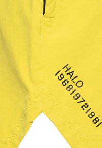 Felgele stof met zichtbare stiksels en zwarte sjabloontekst "HALO 196819721981" diagonaal gedrukt aan de rechter onderzijde.