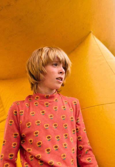 Camisa de manga larga rosa con patrones de girasoles amarillos, llevada por una persona con cabello corto rubio, contra un fondo amarillo brillante.