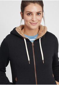 Black hoodie with beige faux fur lining and drawstrings. Features a zip closure and brown faux leather accents near pockets. Striped shirt underneath.