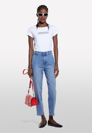 T-shirt bianco con testo stampato, jeans a vita alta azzurri, scarpe nere senza tacco e una piccola borsa rossa. Il modello indossa occhiali da sole bianchi.