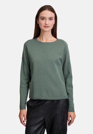 Maglione verde a maglia con scollatura rotonda e spalle cadute, abbinato a pantaloni neri texturizzati. Tessuto morbido e vestibilità rilassata.