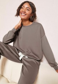 Šedé dlouhé tričko s volnějším střihem, měkkou texturou a bílou mašlí v pase, kombinováno s odpovídajícími kalhotami s volným střihem.