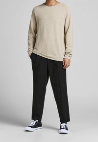 Beige Strickpullover mit langen Ärmeln und Rundhalsausschnitt, kombiniert mit lockeren schwarzen Hosen und schwarzen Turnschuhen mit weißen Schnürsenkeln.