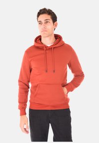 Uomo che indossa un hoodie color ruggine con tasca frontale e pantaloni neri, in piedi con una mano in tasca, mentre guarda di lato.