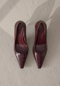 Burgundi színű, hegyes orrú magassarkú cipők, texturált krokodilmintás bőrből, sima belső résszel és finom fényességgel.
