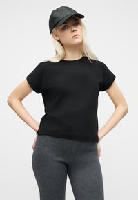 Ξανθιά γυναίκα με μαύρο καπέλο, μαύρο κοντομάνικο μπλουζάκι και γκρι κολάν, στέκεται με τα χέρια στη μέση μπροστά από ένα απλό φόντο.