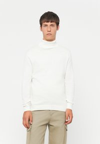 Joven vestindo un suéter de cuello alto blanco y pantalones cargo beige, de pie contra un fondo blanco liso.