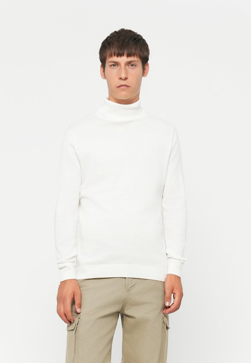 Joven vestindo un suéter de cuello alto blanco y pantalones cargo beige, de pie contra un fondo blanco liso.
