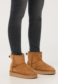 Stivaletti in suede marrone chiaro con punta arrotondata, cuciture a contrasto e fibbie laterali, indossati con jeans neri aderenti.