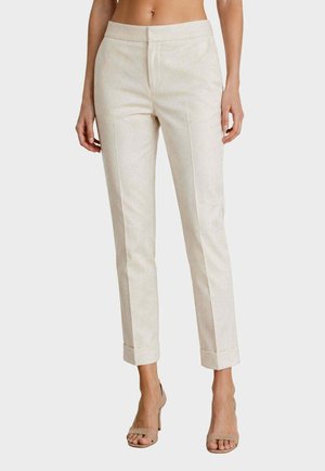 Donna che indossa pantaloni beige chiaro su misura alla caviglia con risvolti e tacchi aperti beige, in piedi davanti a uno sfondo semplice.