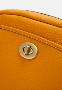 Roh žluté kožené tašky s texturovaným povrchem, vybavený mosazným uzávěrem a kontrastním šitím podél okraje.