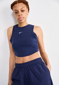 Navy zils apgriezts tanku tops ar ribotu tekstūru un baltu Nike logo. Apvienots ar līdzīgiem navy elastīgiem jostas šortiem.