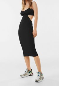 Svart kroppsnära klänning med en utskärning på sidan, tillverkad av ett mjukt tyg. Kombinerad med klumpiga, flerfärgade sportskor.